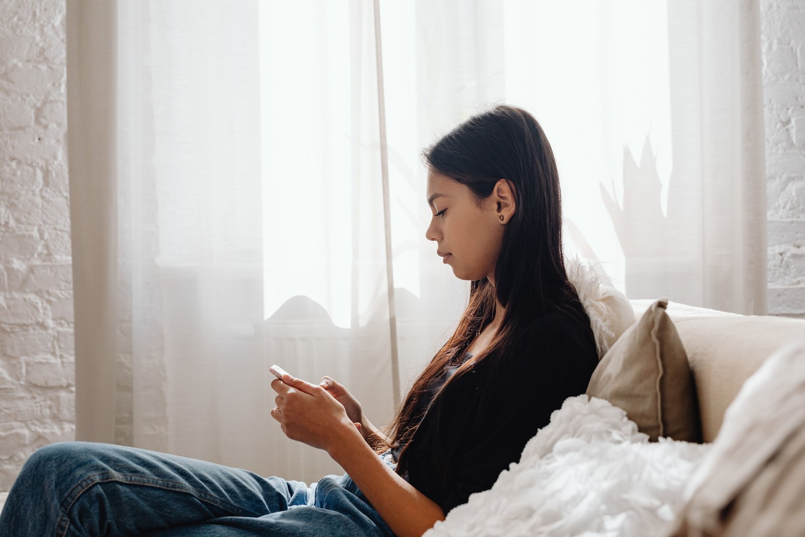 A woman sitting on a bed by a bright window, looking down at her phone
