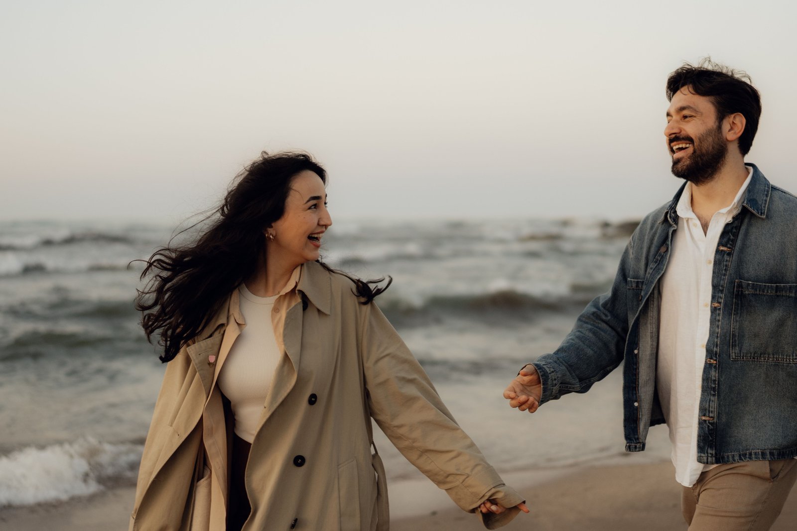 A couple laughing and holding hands while walking along the beach at dusk