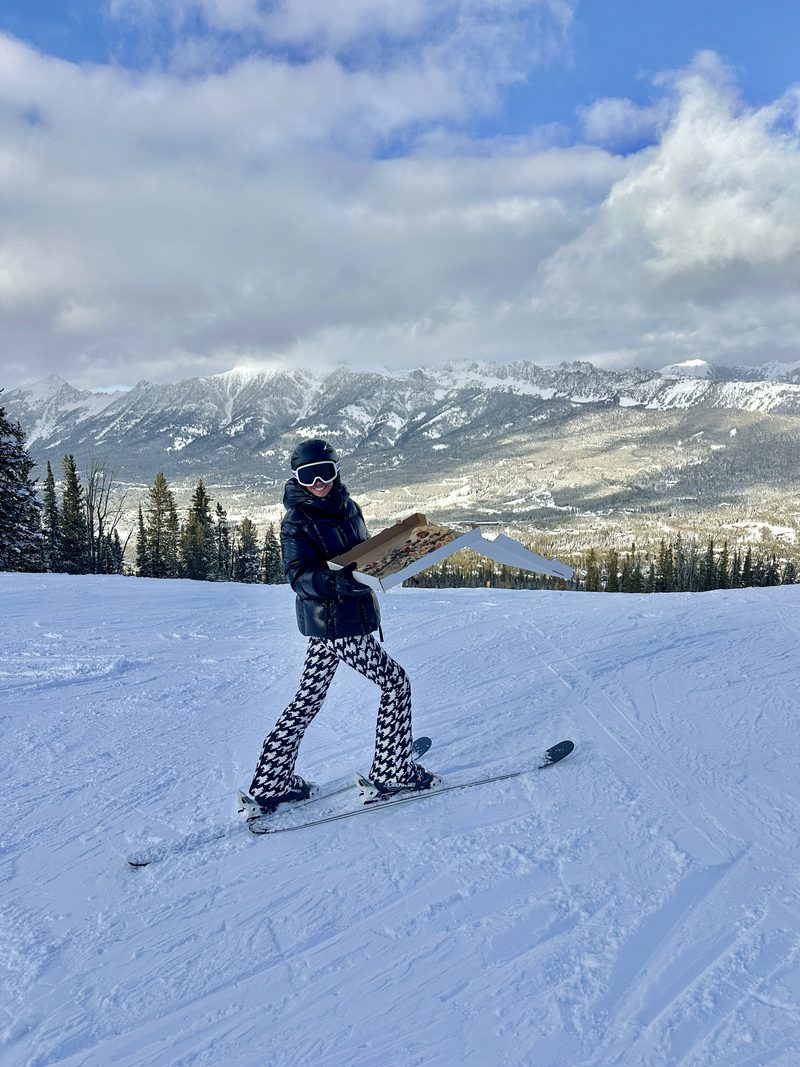 Deryn on skis at the top of a snowy mountain run, holding an open pizza box